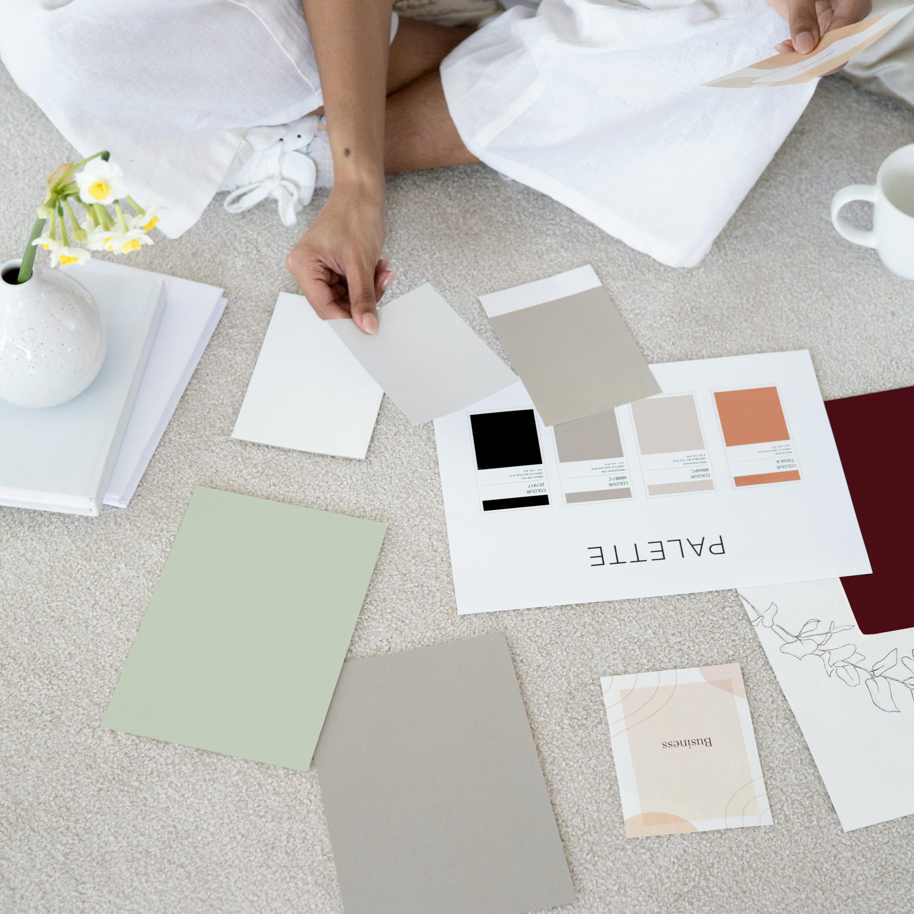 Eine Person sortiert Farb- und Designmuster auf einem Tisch, begleitet von Blumen und einem Notizbuch. Symbol für die Entwicklung einer durchdachten Corporate Identity.