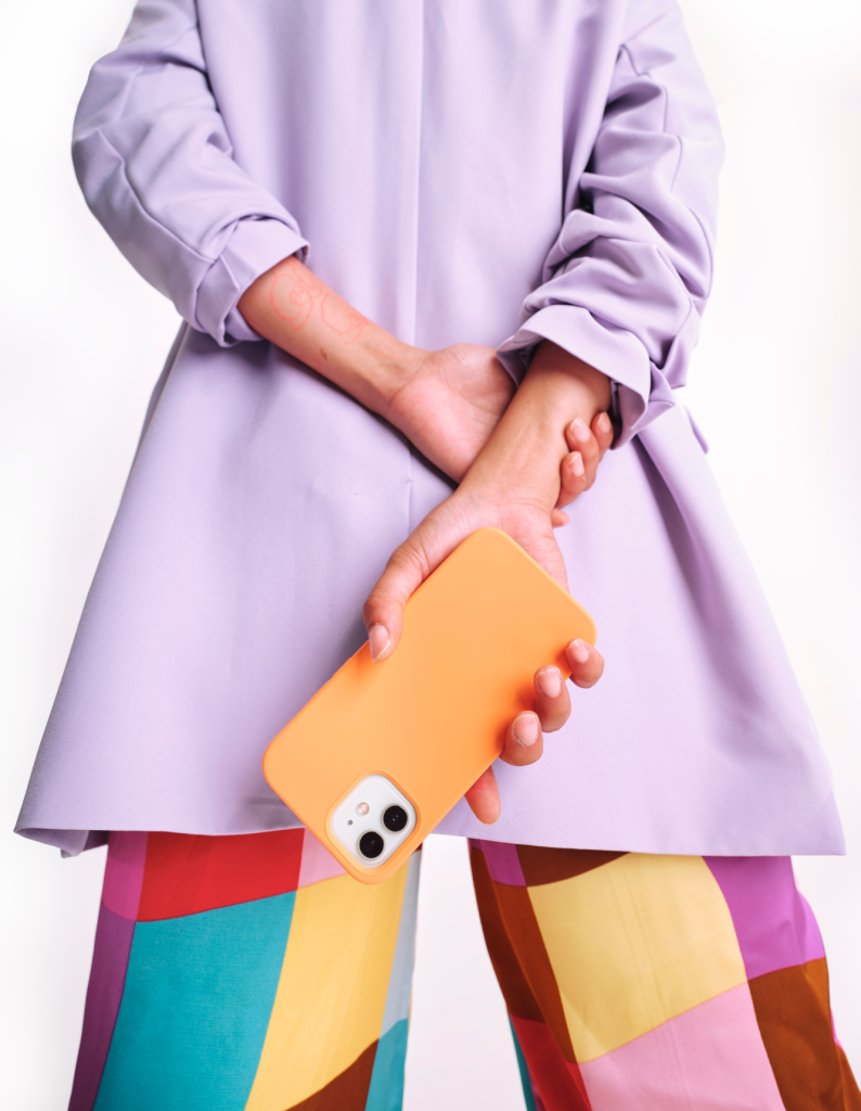 Eine Person in einem lilafarbenen Oberteil und bunter Hose hält ein orangefarbenes Smartphone hinter dem Rücken, als Symbol für modernes Branding.