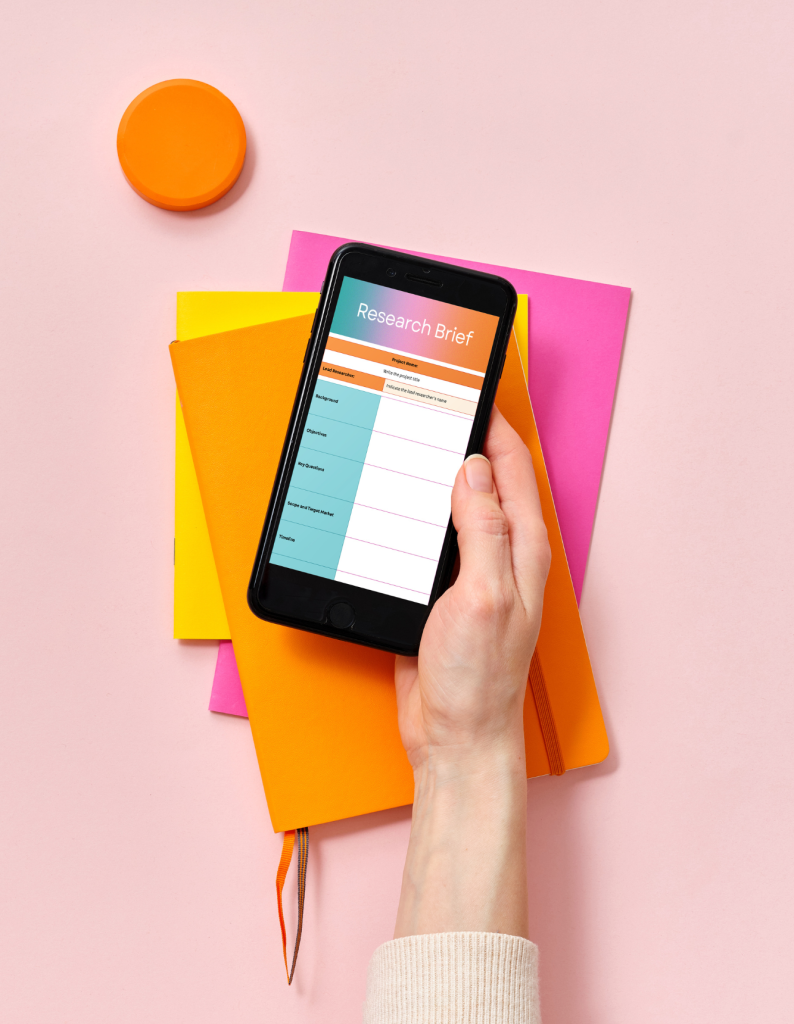 Eine Hand hält ein Smartphone mit ‘Research Brief’ über farbigen Notizbüchern, repräsentierend die Entwicklung einer Corporate Identity.